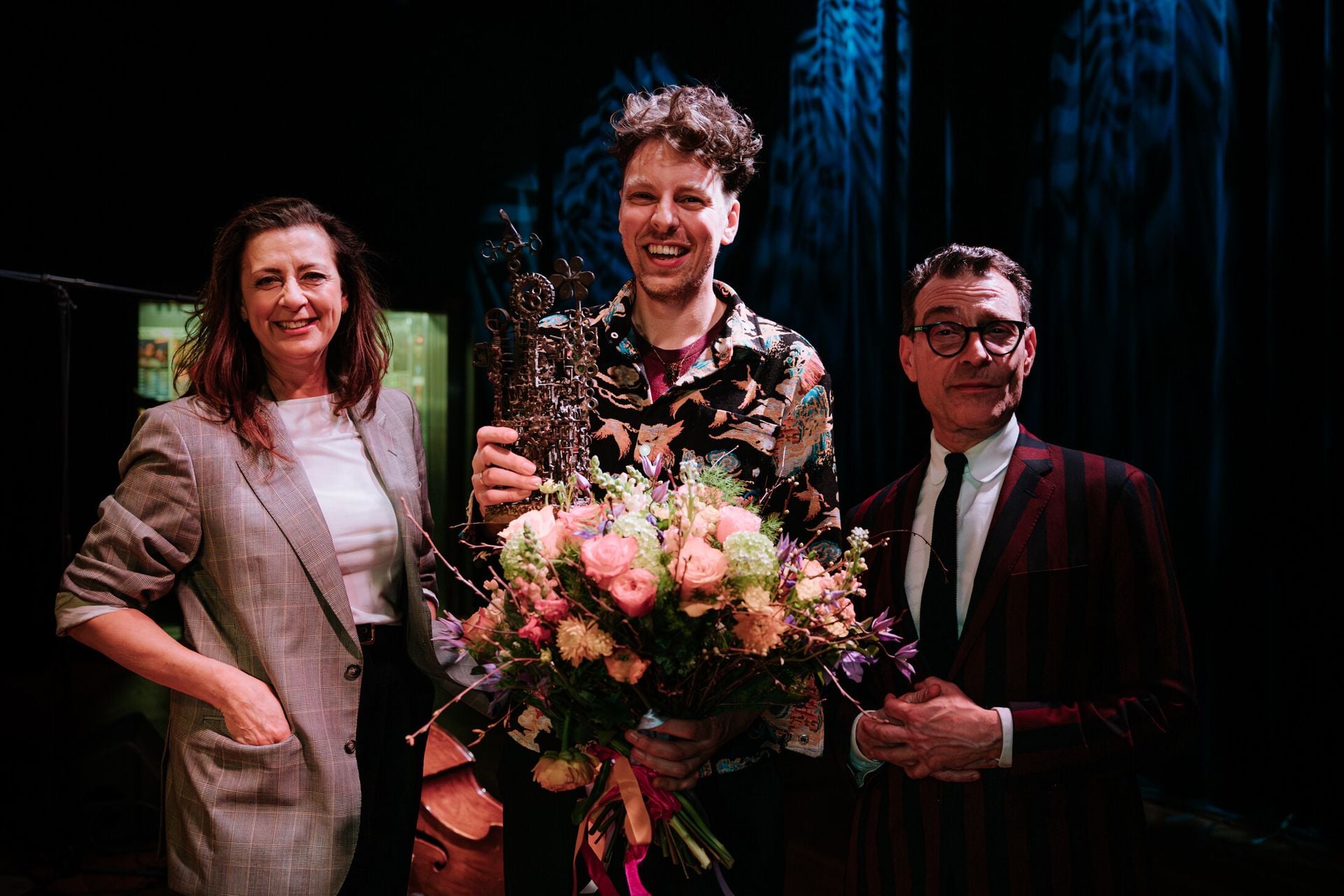 Viktorien van Hulst (Fonds Podiumkunsten), Reinier Baas en oud-winnaar en uitreiker Benjamin Herman op het podium tijdens de uitreiking van de Boy Edgarprijs 2026, met bloemen en een award.