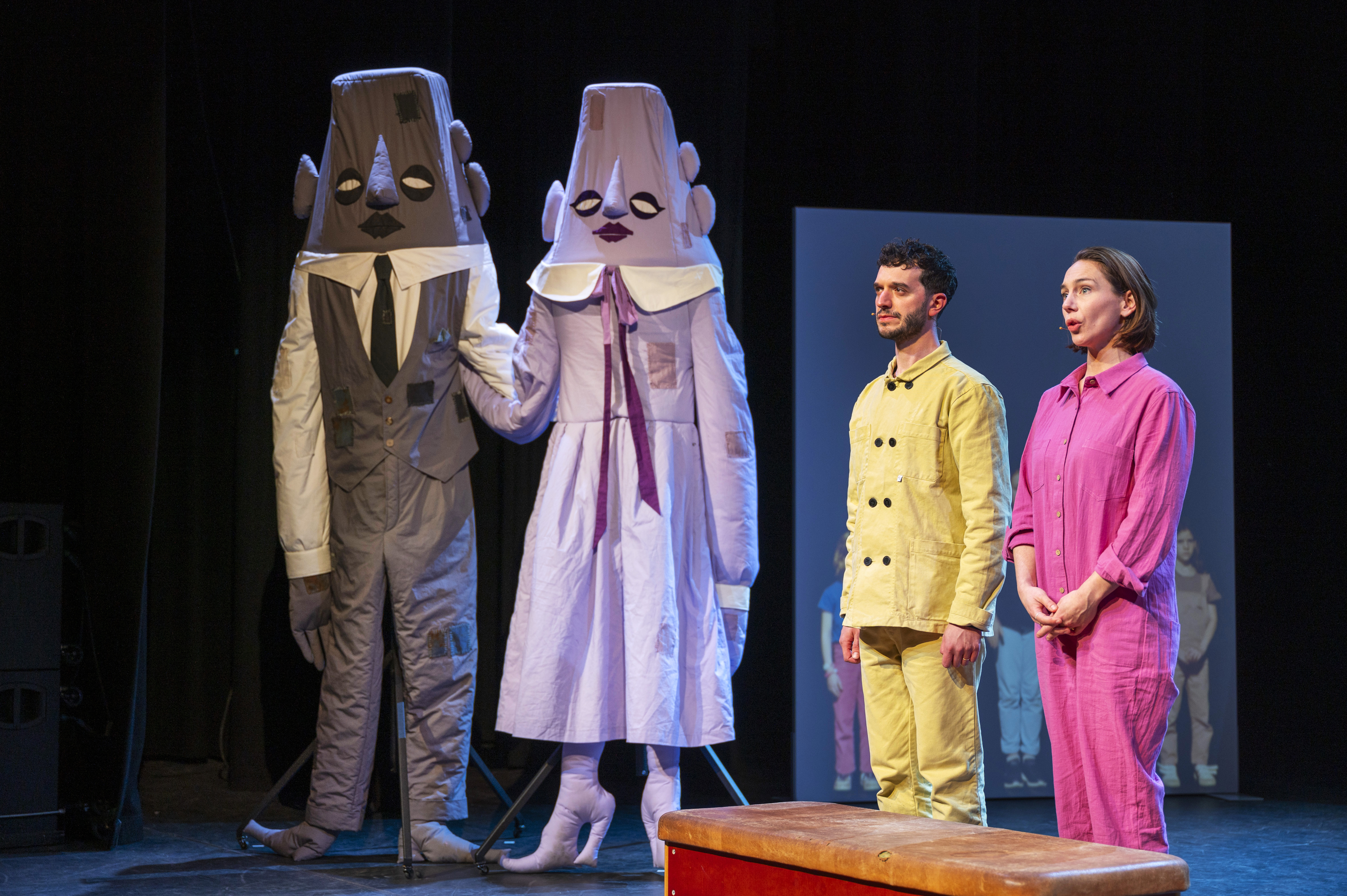 Twee mensen in grote, fantasievolle kostuums staan hand in hand op een podium. Rechts staan twee acteurs in kleurrijke kleding.