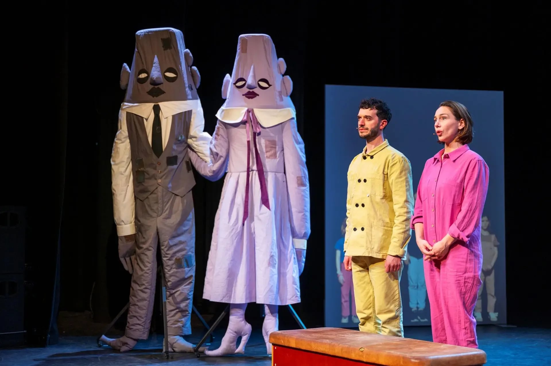 Twee mensen in grote, fantasievolle kostuums staan hand in hand op een podium. Rechts staan twee acteurs in kleurrijke kleding.