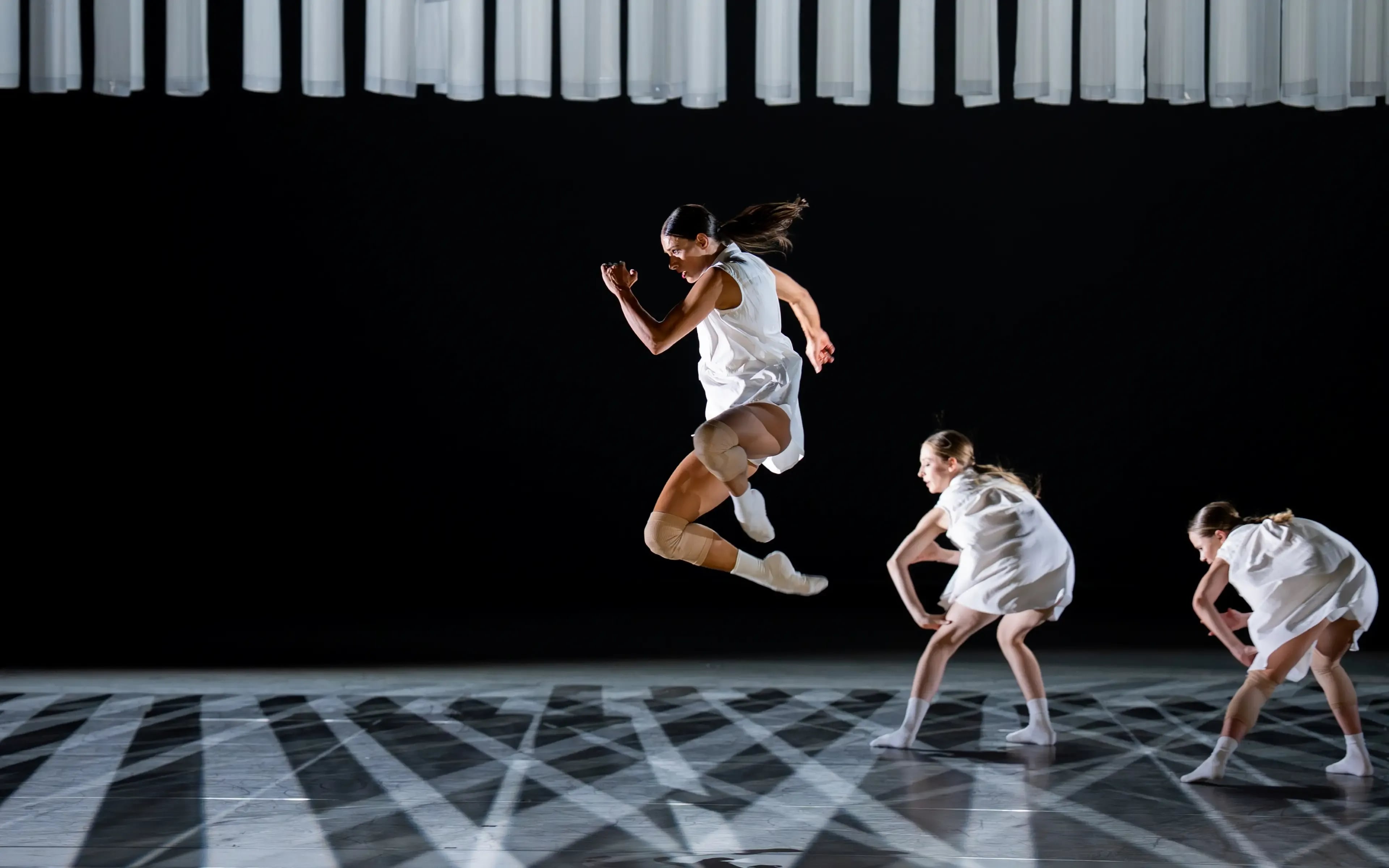 Danser springt hoog in de lucht op een verlicht podium, terwijl twee andere dansers in witte kostuums op de achtergrond bewegen.