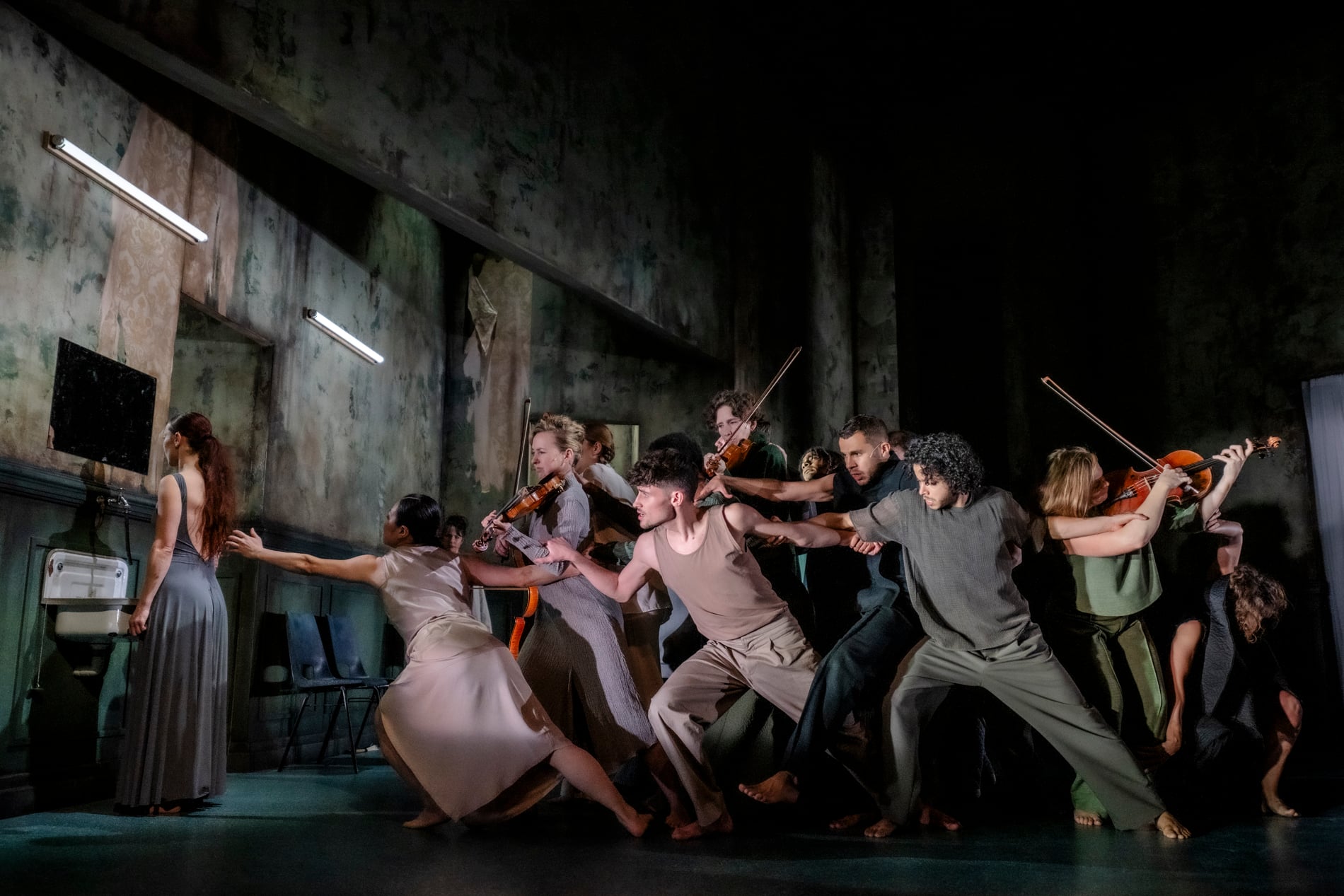 Een grote groep dansers in sobere kleuren kleding rijkt met hun lichamen naar links. Tussen de dansers staan muzikanten die een instrument bespelen.