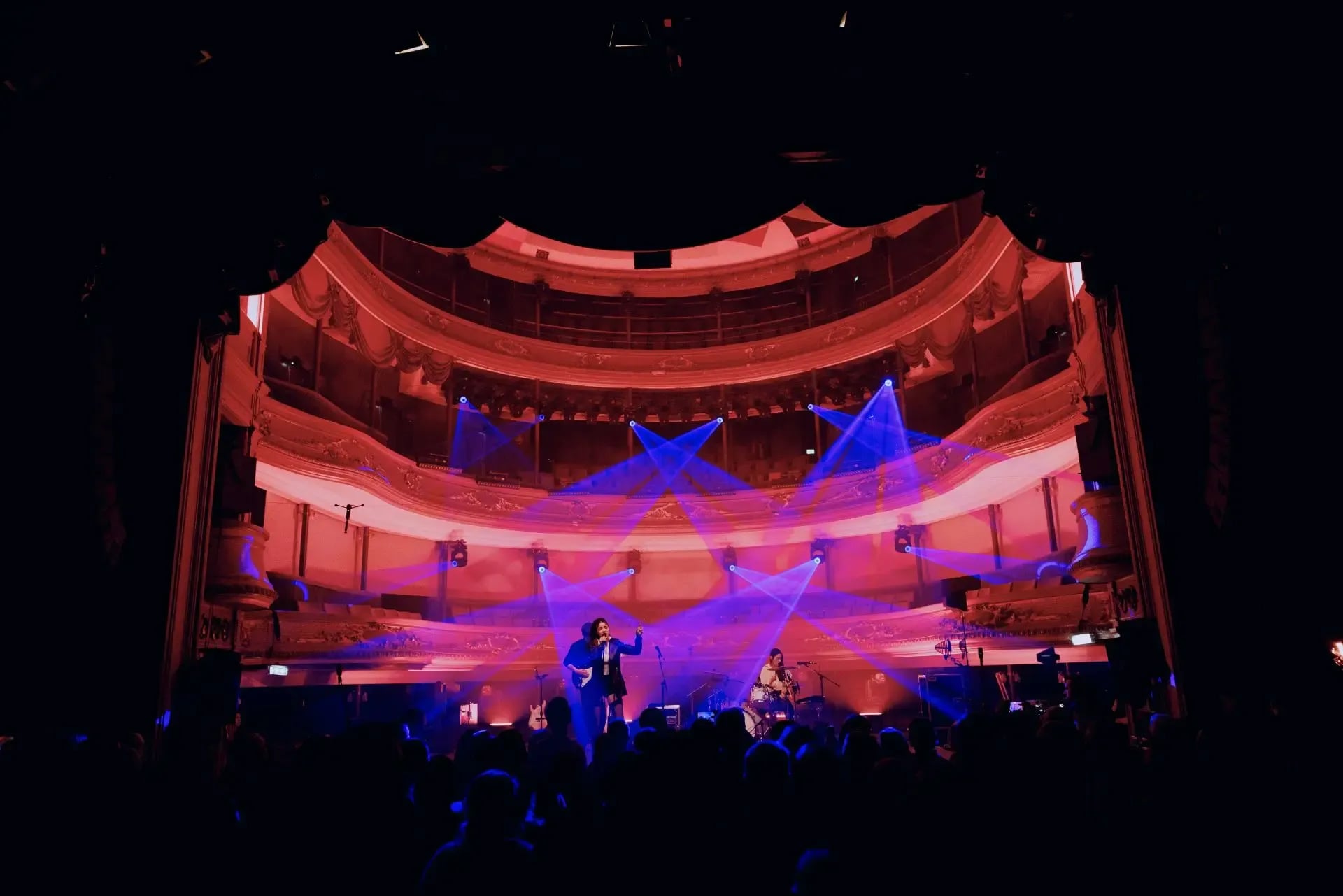 Liveoptreden in een historische theaterzaal met felgekleurde podiumlichten en een groot publiek op de voorgrond