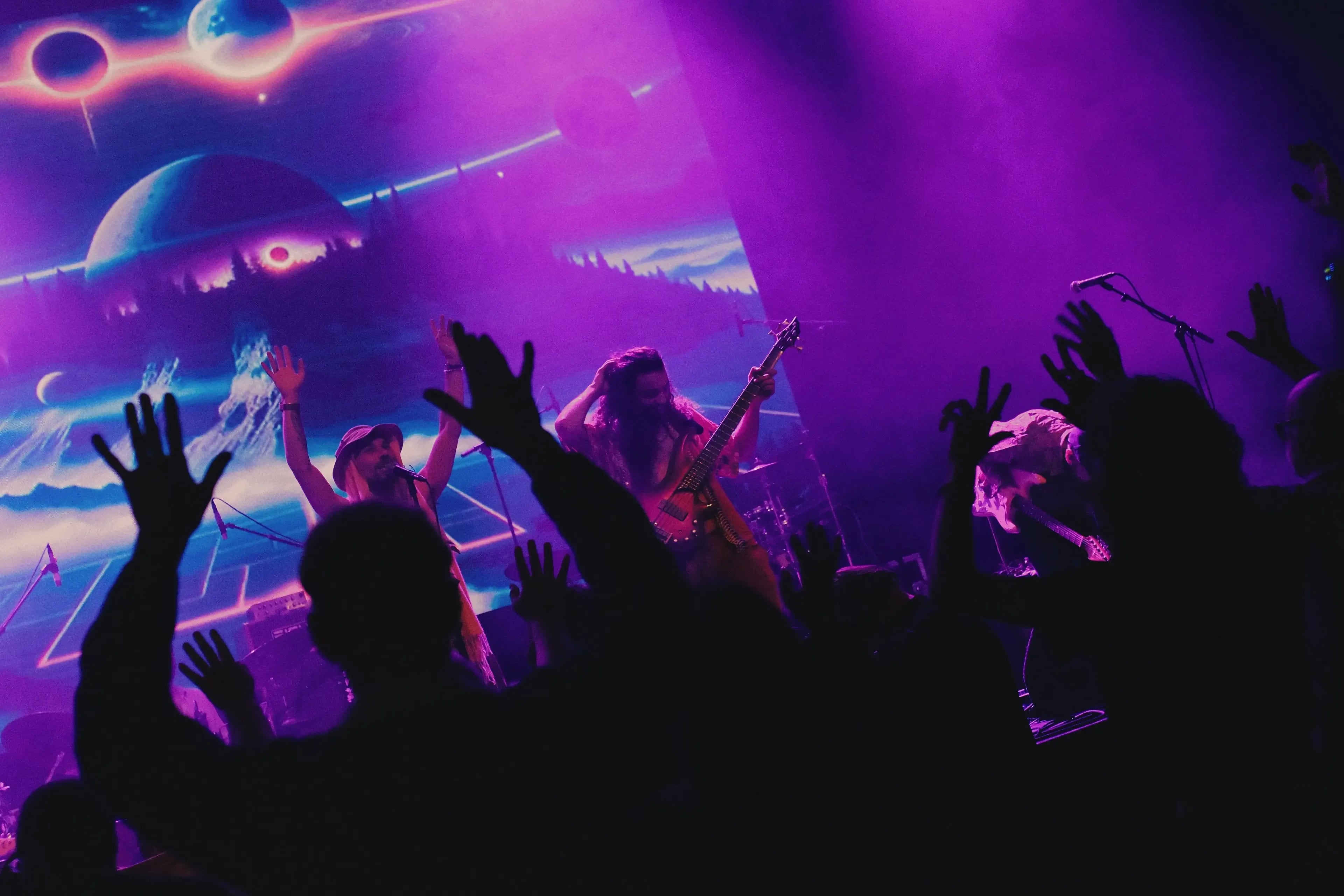A band is performing on a stage with colourful lights while the audience joins in with their hands in the air.