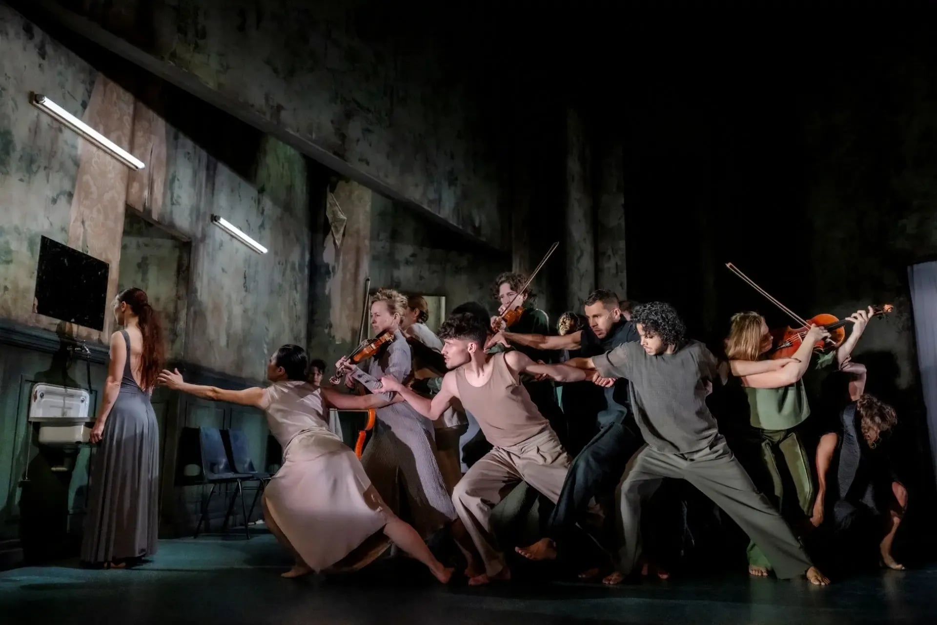 Een grote groep dansers in sobere kleuren kleding rijkt met hun lichamen naar links. Tussen de dansers staan muzikanten die een instrument bespelen.