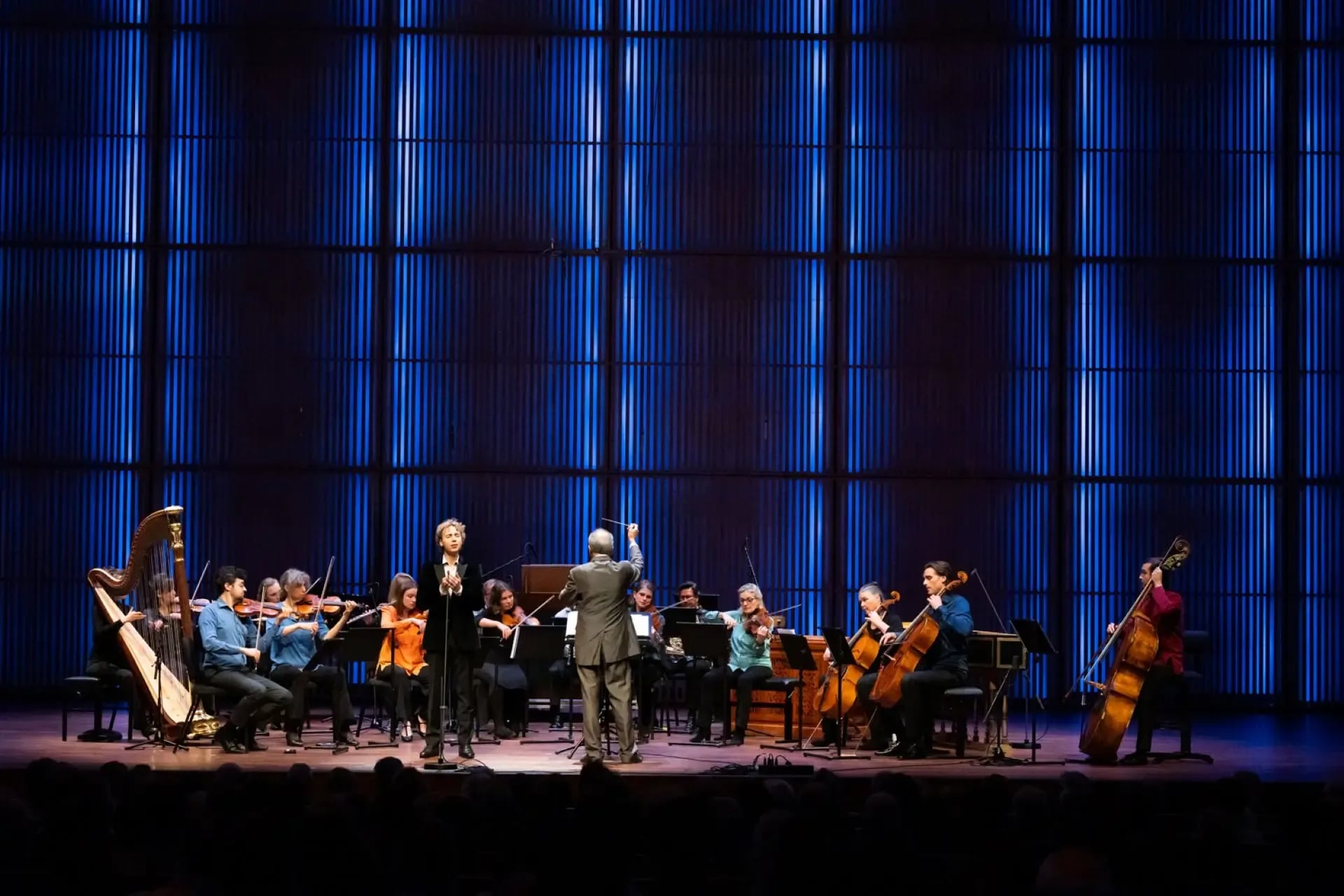 Groot ensemble met strijkers, harp en dirigent op een concertpodium, spelend voor een publiek tegen een wand van verticale blauwe lichtpanelen.