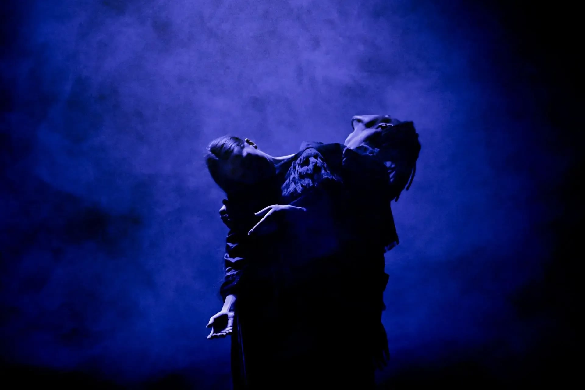Twee figuren in donkere kleding staan dicht tegen elkaar in een blauwe, mistige lichtsetting op een podium.