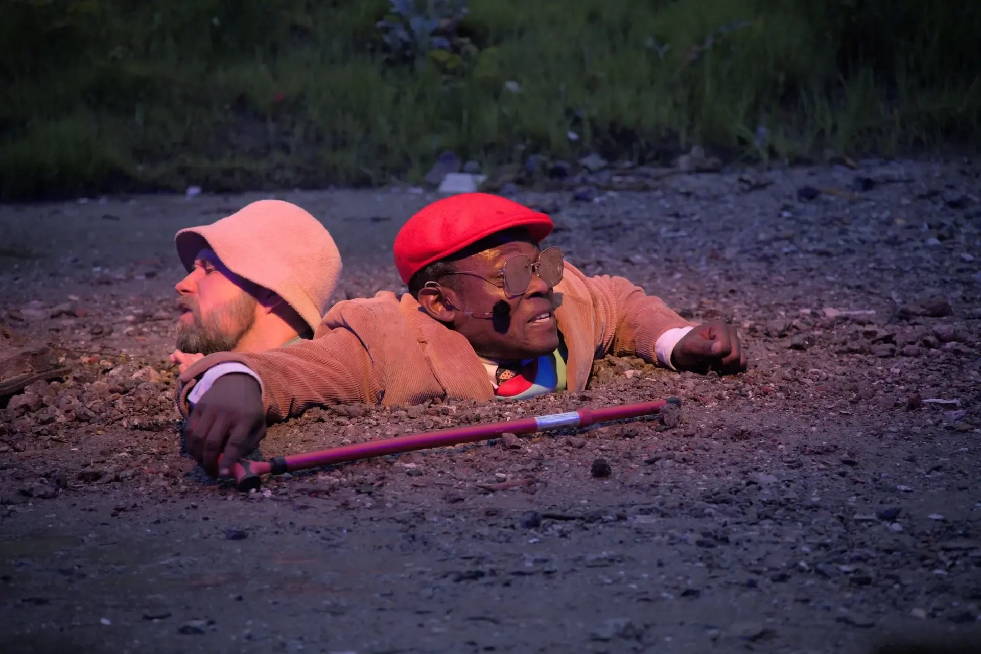 Two men are buried up to their necks in the ground on a gravelly surface; one wears a red cap and holds a cane, while the other wears a light-colored hat and looks away.